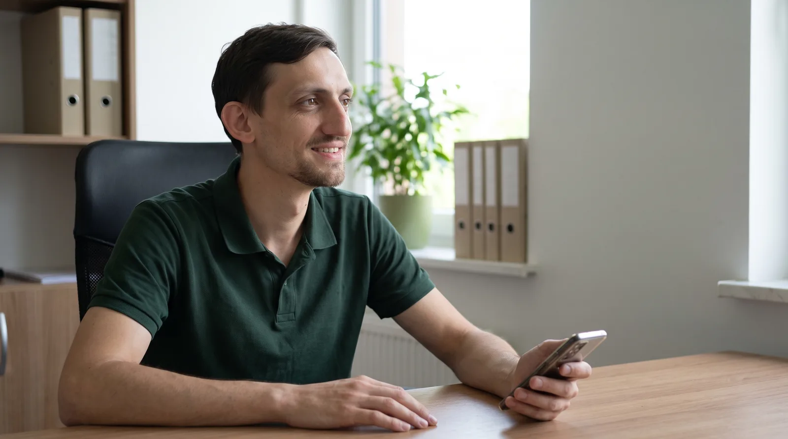 Dawid Jarząbek z Aslandi siedzi przy biurku w biurze z telefonem w dłoni i uśmiecha się — ilustracja AI oparta na jego wizerunku